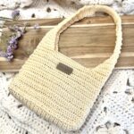 Sac crocheté en fil naturel couleur crème, modèle moderne avec anse épaisse, présenté sur une table en bois avec une décoration florale. Fait main avec une seule pelote.