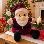 Père Noël au crochet en fil chenille, avec bonnet et tenue bordeaux, barbe blanche, ceinture et bottes noires, assis sur une table.