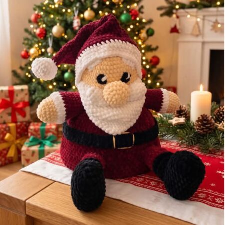 Père Noël au crochet en fil chenille, avec bonnet et tenue bordeaux, barbe blanche, ceinture et bottes noires, assis sur une table.