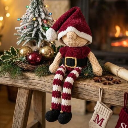 Lutin de Noël au crochet avec bonnet bordeaux, jambes rayées crème et bordeaux, chaussures noires et petites cloches dorées.